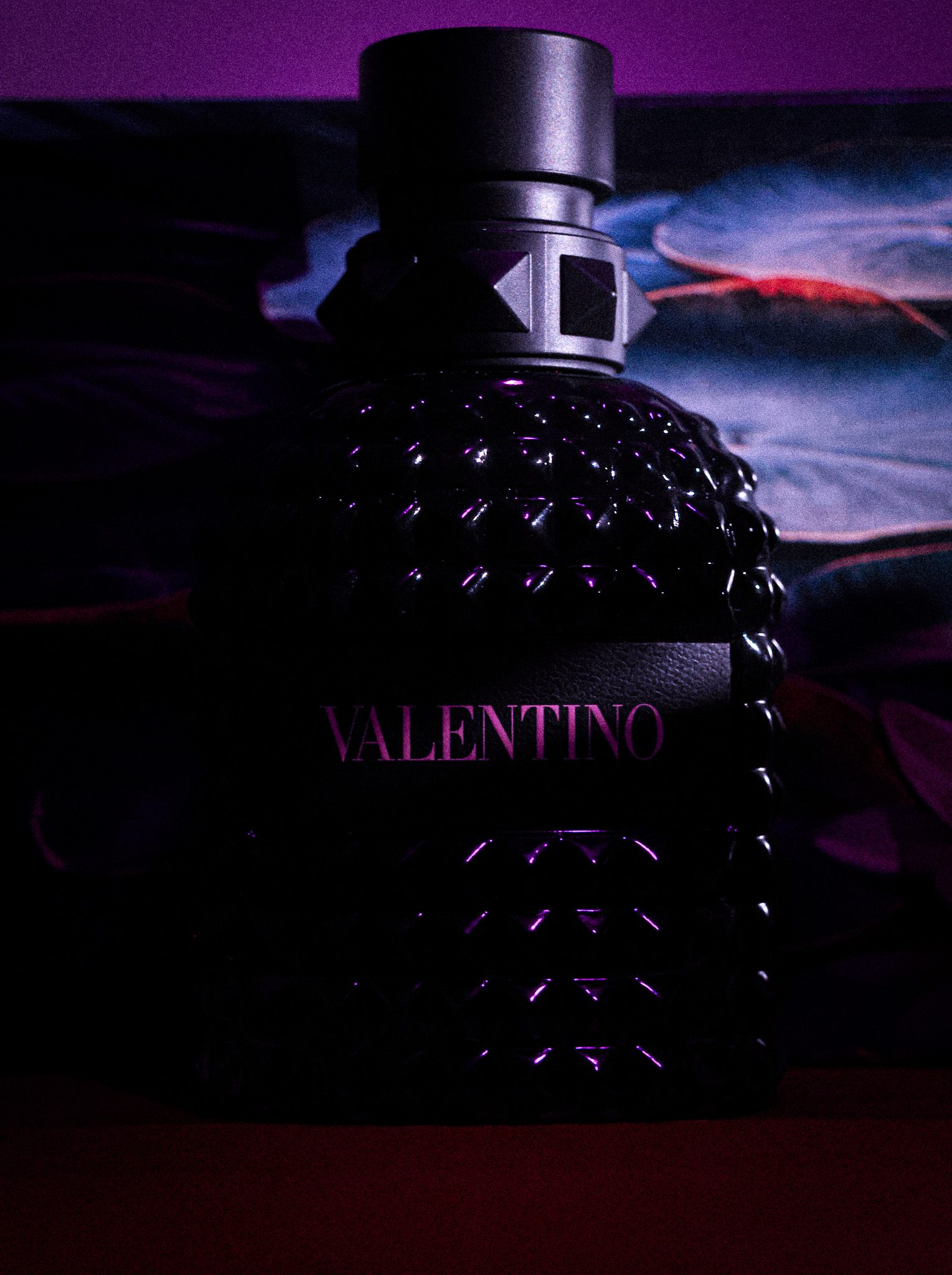 A Valentino cologne bottle lit in moody purple against a dark backdrop.
