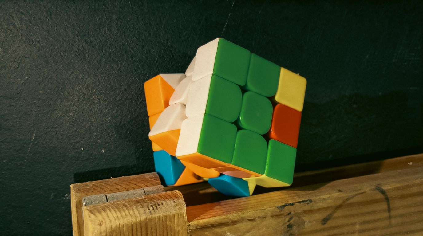 A colourful Rubik's Cube mid-turn, balanced on a wooden edge against a dark green wall.