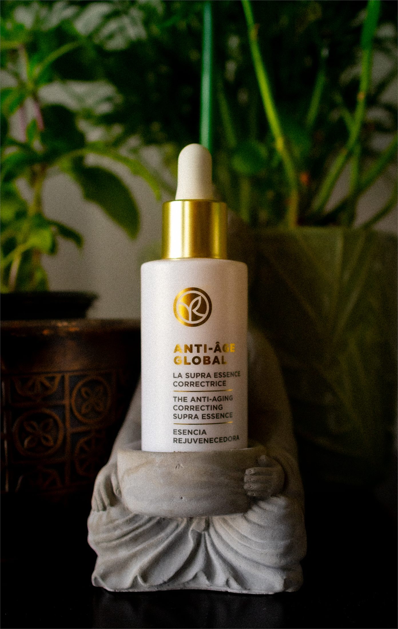 An Yves Rocher Anti-Age Global essence dropper bottle held by a small concrete monk figurine, greenery behind.