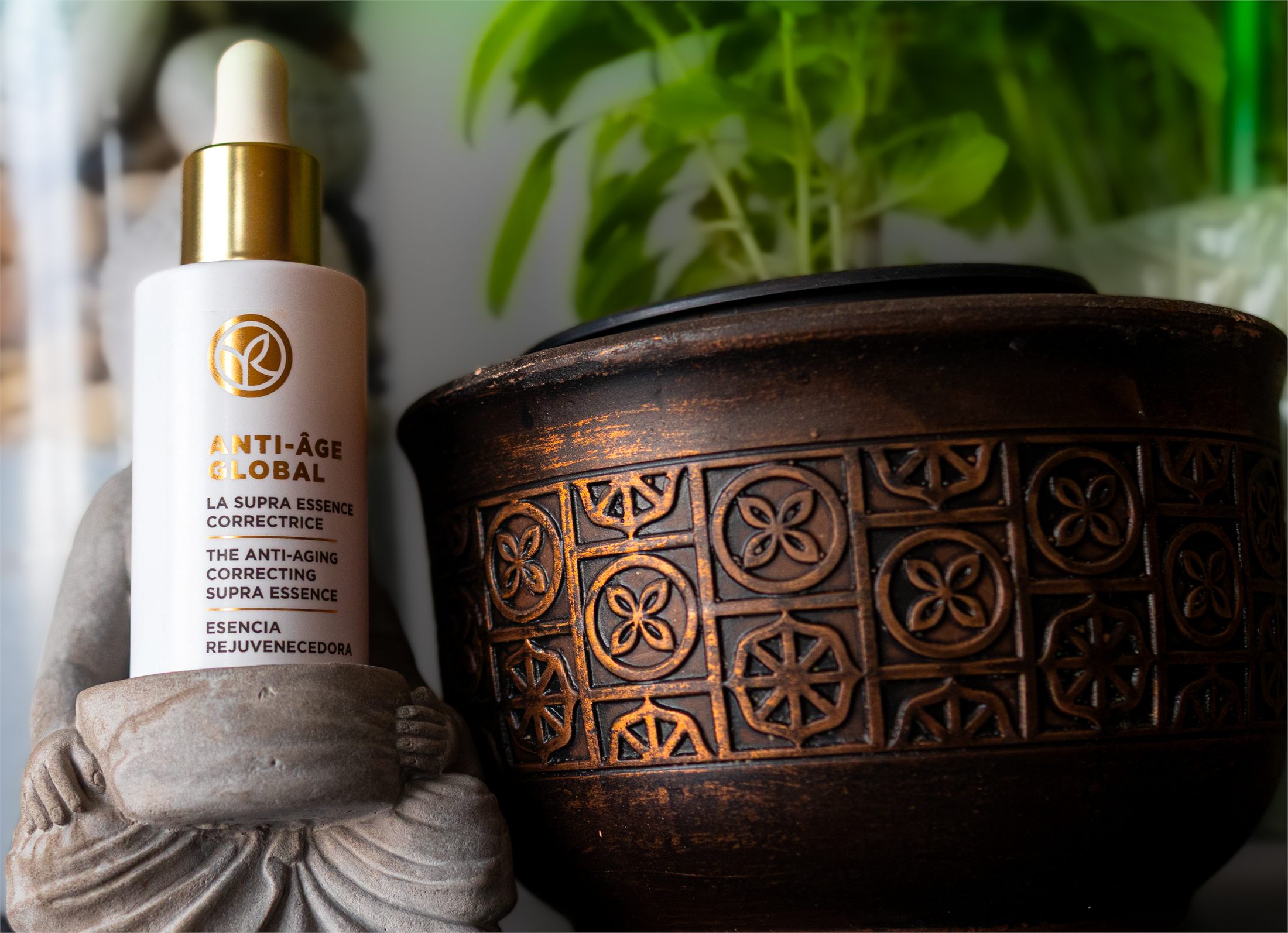 The Yves Rocher essence bottle held by the monk figurine beside a patterned copper pot and a plant.
